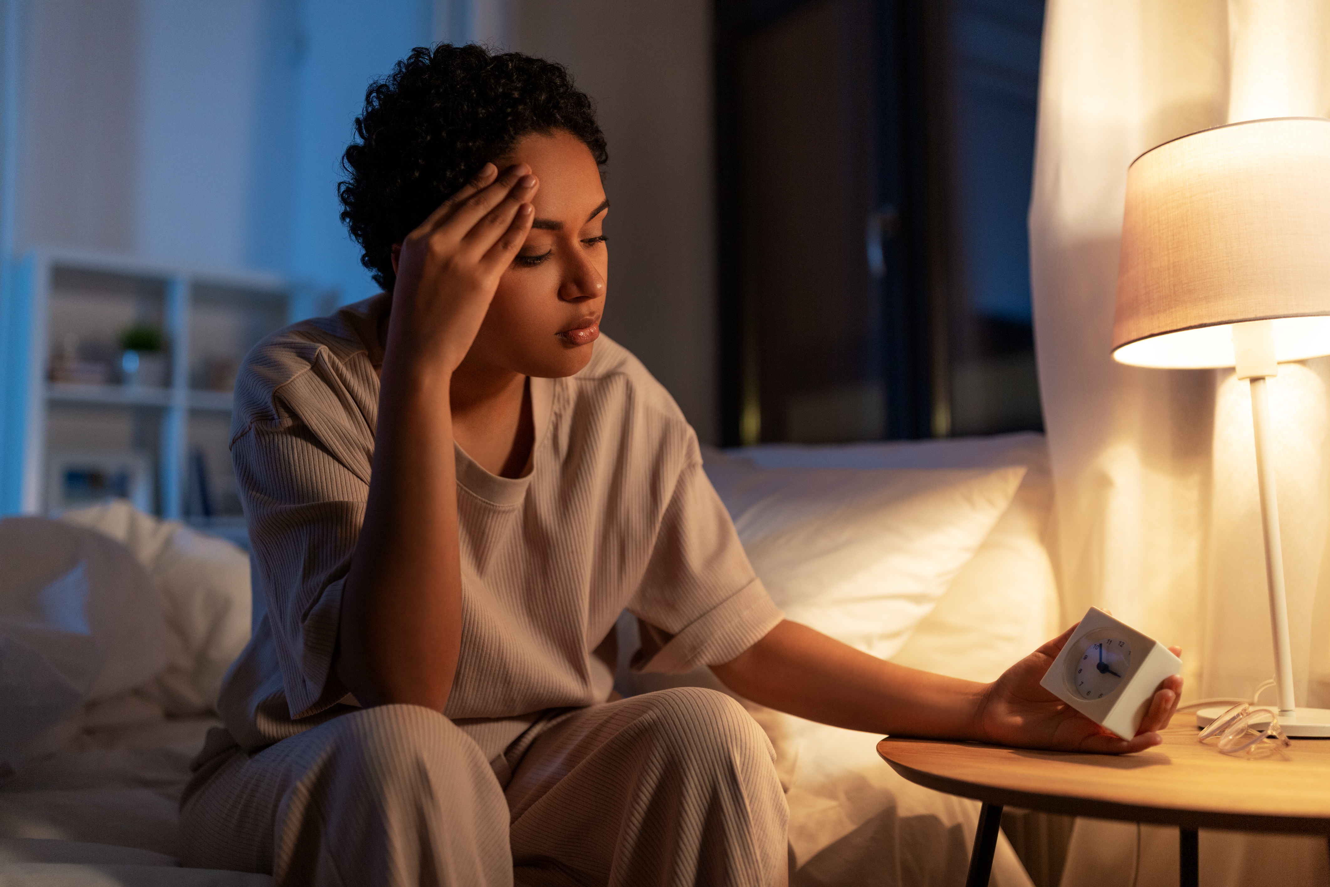 woman sitting on the edge of her bed at night with a hand to her aching head, holding an alarm clock because she can't sleep - insomnia and sleep disruption during menopause